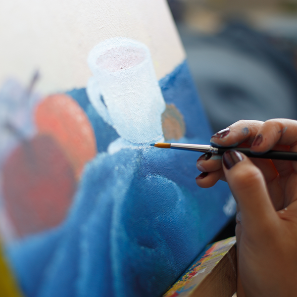 Someone painting a still life of fruit, a cup, and table cloth on a canvas.