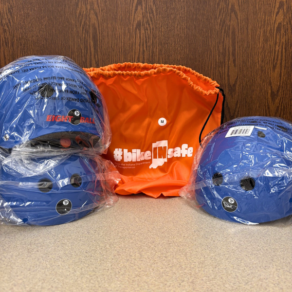 Youth-sized bike helmets in plastic wrap are on either side of an orange bag with #bikeINsafe on the front.