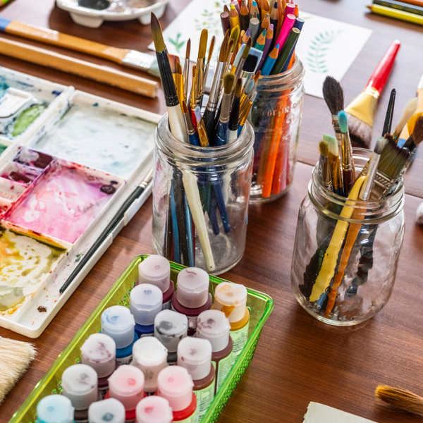 Various art supplies, like paints and paint brushes in jars, on a table.