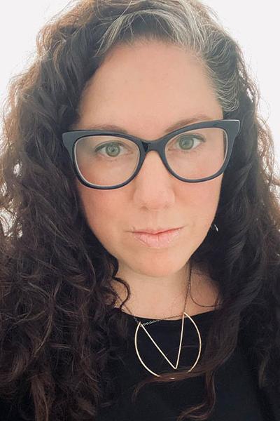 A close-up of a woman with large eyeglasses and wearing a V-shaped necklace over a dark-colored blouse.