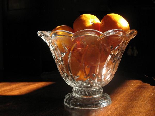 A decorative glass bowl filled with several bright oranges sits on a wooden table. Sunlight highlights the fruits and creates reflections on the intricately patterned glass. The background is dark, contrasting with the illuminated bowl and oranges.