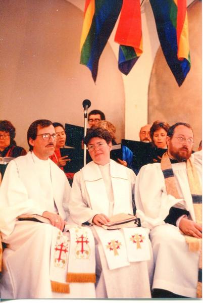 Three clergy, two men and a woman, dressed in white robes, sit together in a church. Members of the church choir can be seen seated behind them, and above are three rainbow banners hanging from the ceiling.