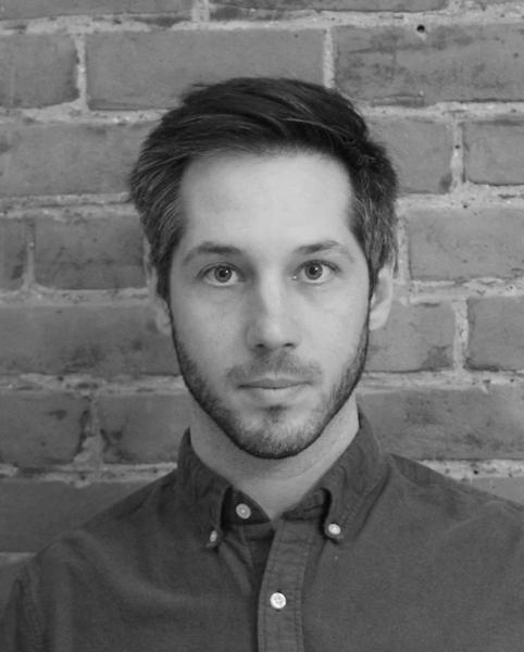 A black-and-white portrait of a bearded man in front of a brick wall