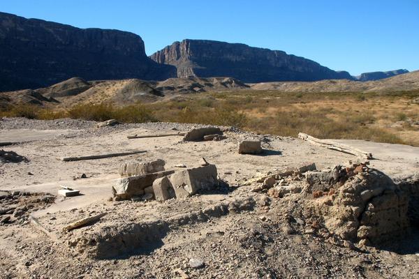 A desolate landscape includes scattered concrete ruins in the foreground. The background features arid terrain and rugged, steep cliffs with a large gap in the ridgeline. The entire scene is under a clear blue sky, with sparse vegetation dotting the expansive landscape
typical of Big Bend National Park.of Big Bend National Park.
