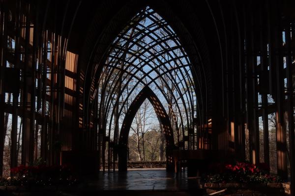 A view from inside a chapel with an arched wooden framework and glass walls, looking out toward leafless trees and greenery. Sunlight highlights parts of the interior, and red poinsettia flowers are visible at the edges.