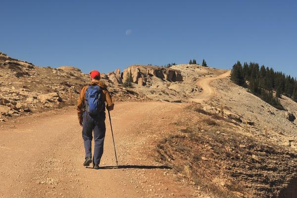 A hiker wearing a red cap and backpack walks with a trekking pole on a dirt road to the Medicine Wheel in Wyoming. The road winds through a dry, rocky landscape with sparse vegetation under a clear blue sky. The moon hangs faintly in the sky above the horizon.