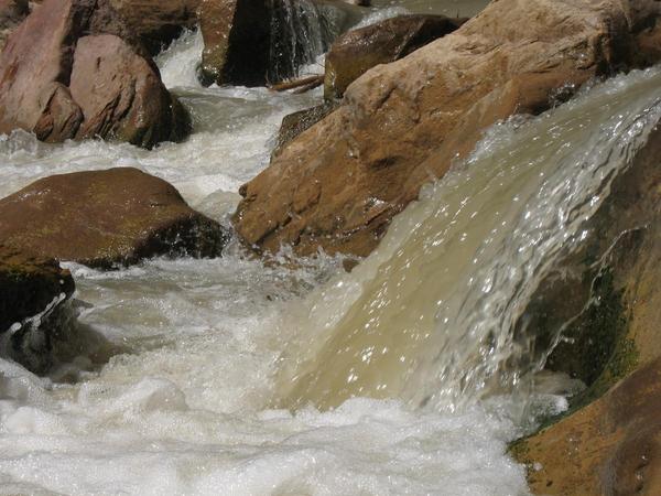 Muddy water flows over rocks in a stream, creating white foam and splashes. Large rocks line the sides as water cascades down a small drop. The scene suggests movement and natural erosion.