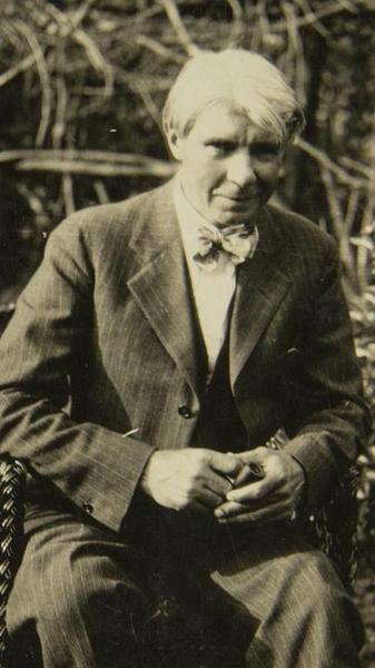 A black-and-white outdoor portrait of a seated man in a suit with bow tie. His hair is white and his elbows rest on the arms of the chair with his hands holding a pipe above his lap.