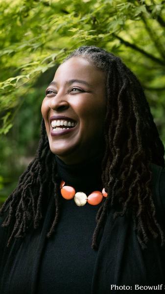 Portrait of a African American woman smiling broadly with a colorful necklace and standing in front of a background of green foliage.