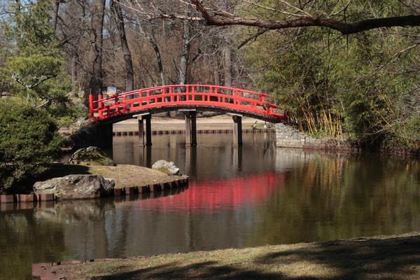 A bright red bridge spanning serene still waters showing the bridge's reflection and flanked by trees at both ends of the bridge.