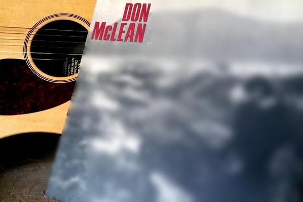 A vinyl record cover with the words Don McLean written on it leans against an acoustic guitar. The album cover showcases a blurred black and white image of a figure standing outdoors.