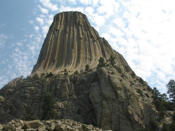 A large, imposing rock formation rises against a partly cloudy sky. The columnar structure exhibits vertical striations and is surrounded by rocky terrain with scattered trees at its base.