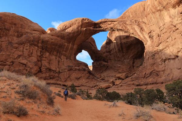 Large stone arch with small hiker in the foreground and a blue sky in the background.