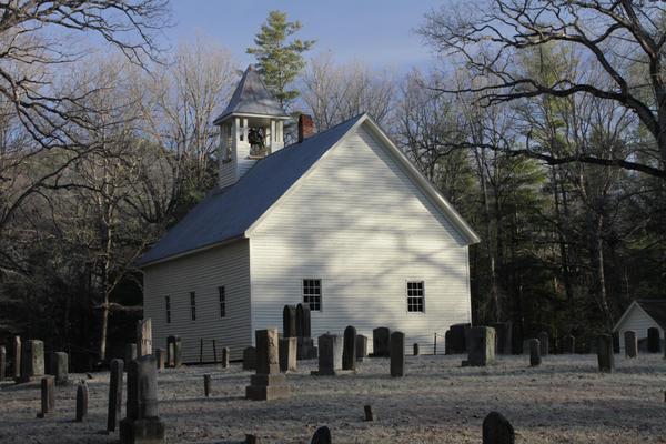  A white wooden church with a small bell tower is surrounded by old gravestones in a cemetery, with leafless trees and pine trees in the background under a clear sky.