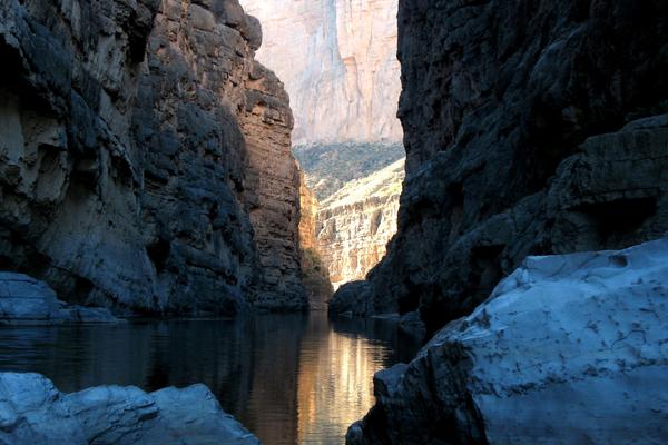 A narrow canyon in Big Bend National Park features tall, rugged rock walls with a calm river flowing through it. Sunlight illuminates the cliffs in the background, creating a stark contrast with the shadowed areas in the foreground. Large rocks are visible in the river,
enhancing the majestic beauty of the scene.