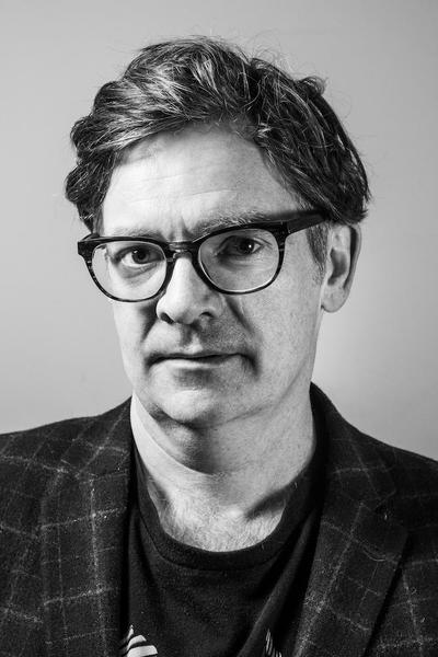 A black-and-white portrait of a man with thick, dark hair and large glasses.