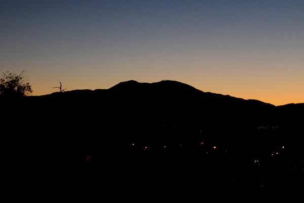 A silhouette of Saddleback Mountain against a pre-dawn sky, with a gradient from deep blue to orange. Distant city lights are scattered at the base of the mountain, creating a serene and tranquil atmosphere.