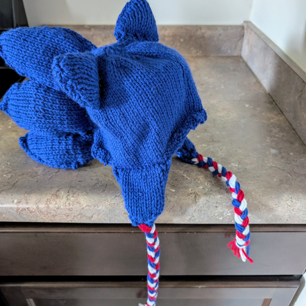 A blue knitted hat resembling a hedgehog with pointy ears and quills, placed on a countertop. It has red, white, and blue braided ties, giving a playful tone.