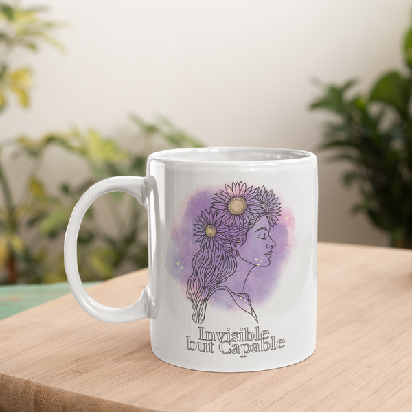 A white mug on a wooden surface features an illustration of a woman with flowers in her hair, set against a purple background. Text reads "Invisible but Capable." Lush green plants in the blurred background add a calming atmosphere.