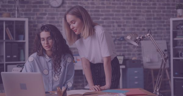 Two women focus on a laptop in an office with a brick wall. One stands leaning over the desk, and the other sits, creating a collaborative atmosphere.