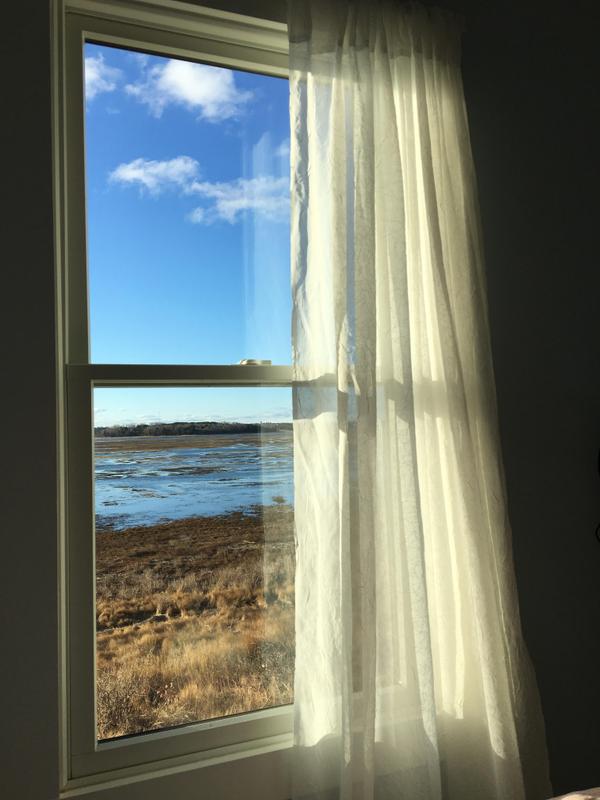 A window with a billowing curtain looks out
on a winter marsh and bright blue sky, with the high tide water from the sea starting to cover the marsh grasses.