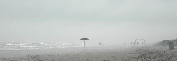 A beach umbrella stands in the fog, tiny figures beyond in the distance.