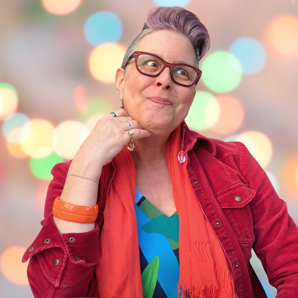 Picture of Lisa with pink hair in a high wave, wearing red glasses, a turquoise and green patterned shirt, red corduroy jacket, orange scarf and orange bracelet with dangly handmade earrings, and a silly look on her face