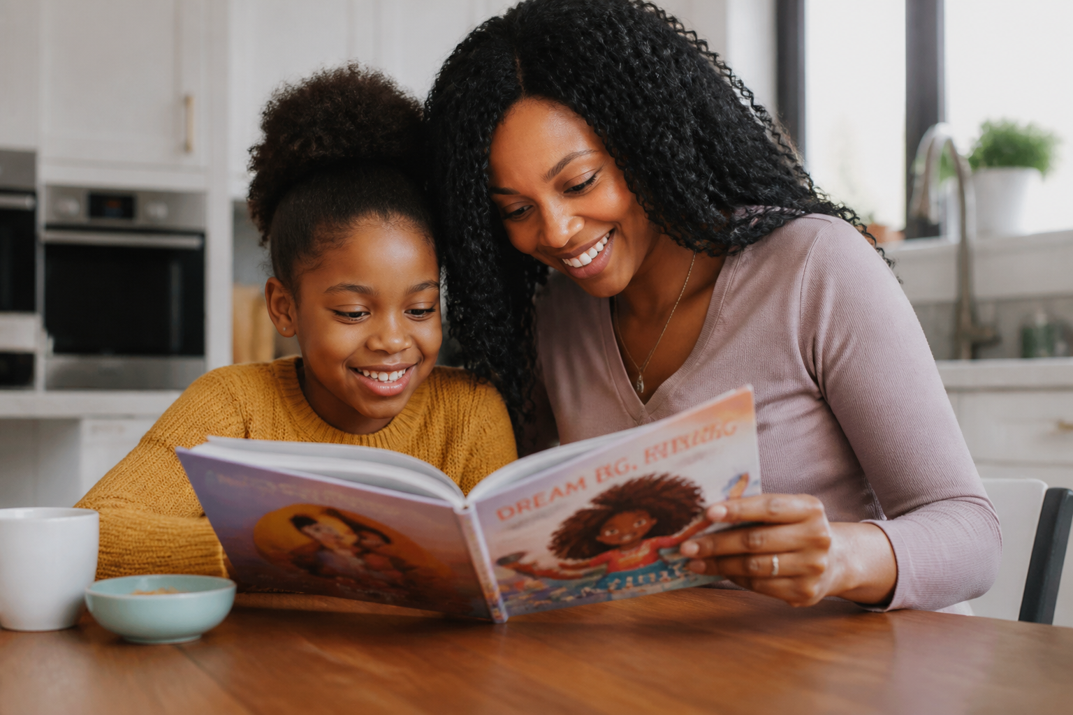 A parent and their smiling child reading a book together at a sunlit kitchen table