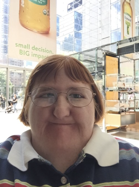 A selfie of a woman with short red hair and glasses in front of what appears to be a mall in the US.