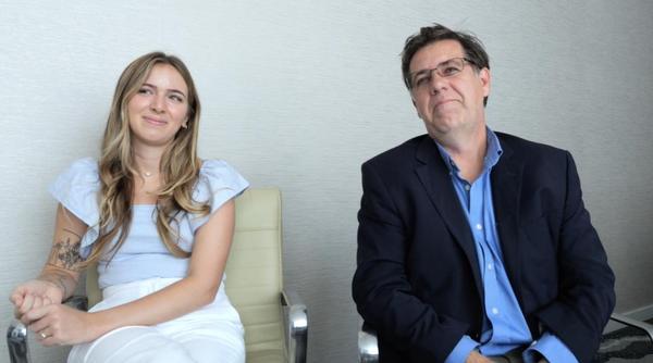 Two people are sitting in beige office chairs in front of a white wall. Dr. Gensemer is a woman with long, wavy blonde hair. She wears a blue shirt and
white pants and has flower tattoos on her arm. Next to her sits Dr. Norris, a man with short brown hair, grey glasses, a blue shirt and a dark blue jacket on top. 
