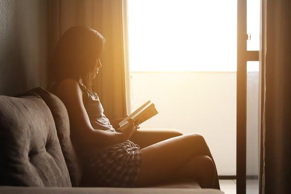 A person sitting on a couch reading a book while the sun is shining inside from the large windows. 