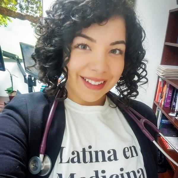 A Latinx woman with curly black hair wearing a stethoscope around her neck. She wears a shirt saying "Latina en Medicina".