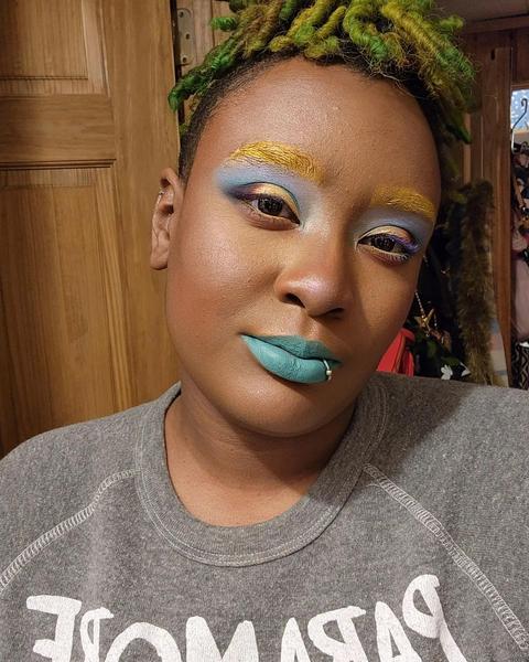 A black woman with green hair and colorful makeup: She has yellow eyebrows, blue and gold eyeshadow and turquoise lipstick. 