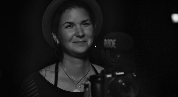 A woman with short brown hair and a grey hat sits behind a camera with a shotgun microphone attached.