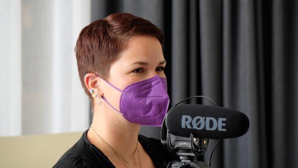 A woman with short brown hair and a purple face mask sits on a  beige chair behind a black camera with an attached shotgun microphone. 