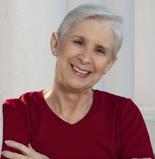 A woman with short, gray hair standing in front of a white wall. She wears a red shirt and smiles brightly. 