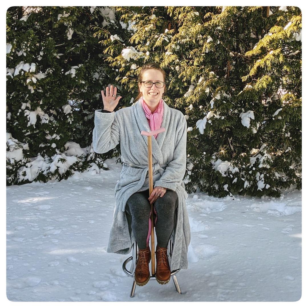Brianne, a white woman in her mid-30s wearing black framed glasses and dark brown hair in a messy bun, sits perched on a high stool in a snowy yard holding a
cane between her thighs while smiling at the camera with evergreen trees in the background. She’s wearing a light blue bathrobe and a pink scarf, and holds one bare hand up to wave at the camera