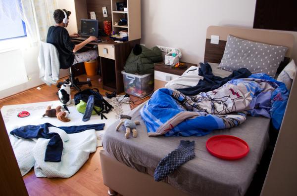 Teen boy wearing headphones sitting in a messy bedroom.
