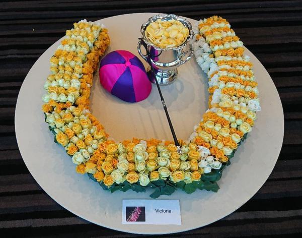 A horseshoe of mixed yellow roses with jockey's cap and crop and silver trophy filled with yellow rose petals