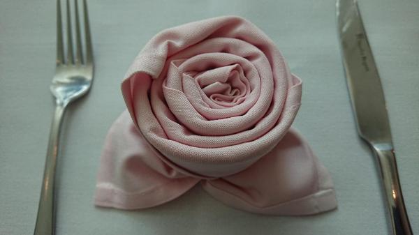 A pale pink dinner napkin folded into the shape of a rose with silver knife and fork