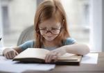Little girl with big glasses sitting at a desk reading a book.