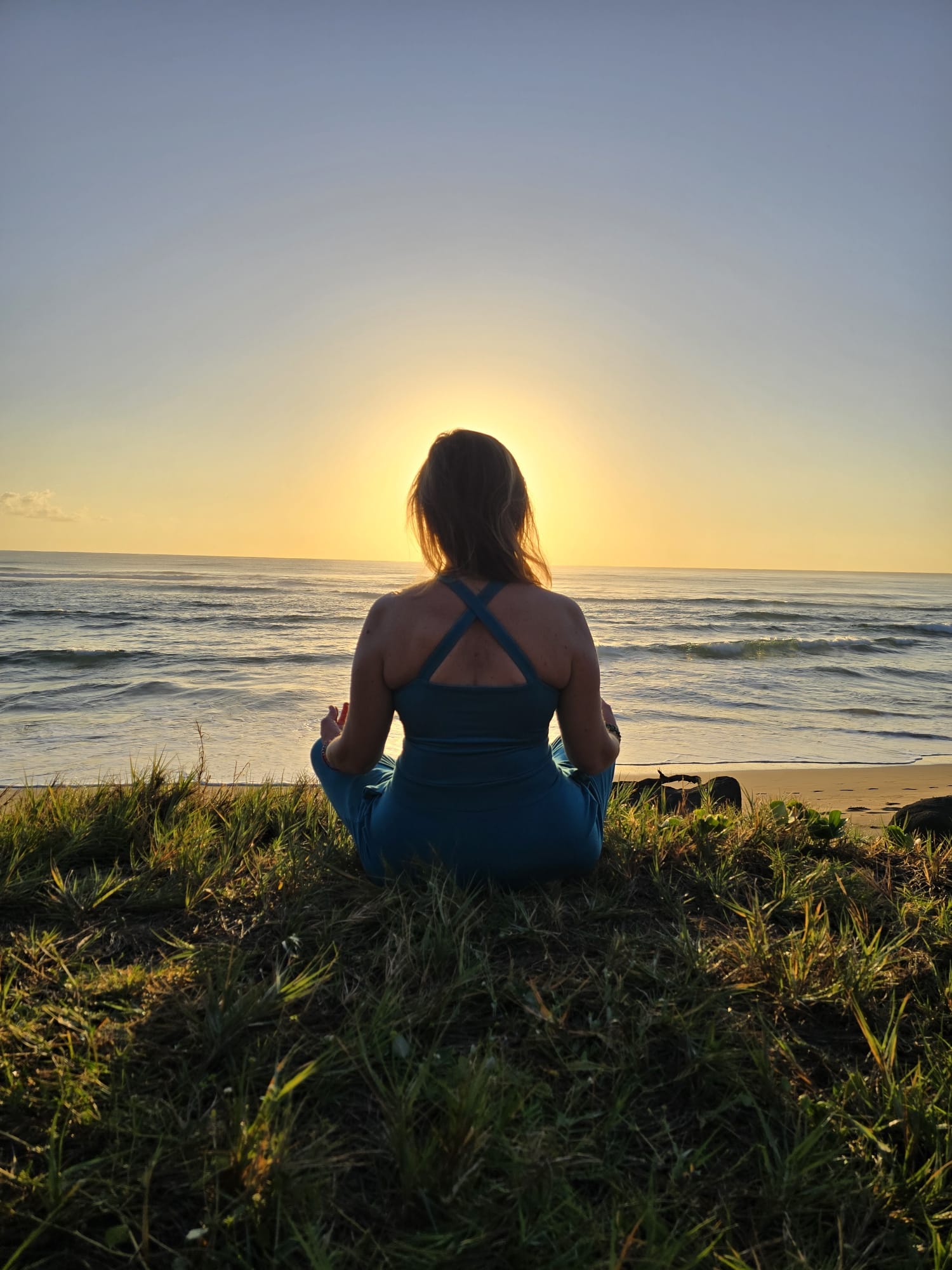Anya Love meditating cross-legged on the grassy oceanfront lawn at Kauai Shores as the sun rises over the water