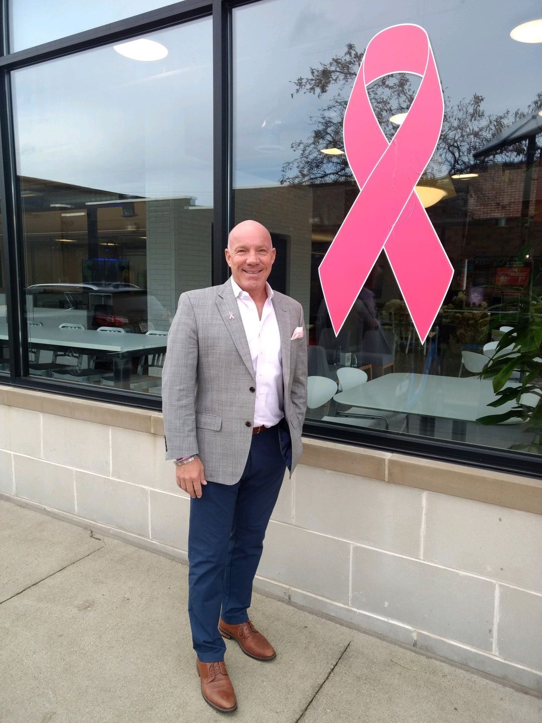 Michael Pesendorfer smiling in a grey blazer beside a large pink breast cancer awareness ribbon