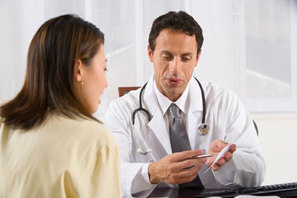 A male doctor with a white coat and a stethoscope around his neck talks to a female patient with brown hair waring a beige shirt.