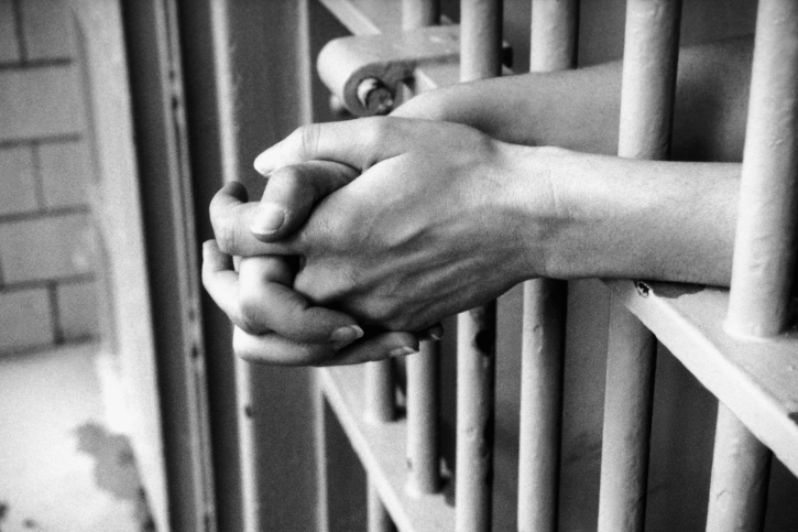 A black and white image of the bars of a jail cell. A pair of hands are clasped and resting between the bars.